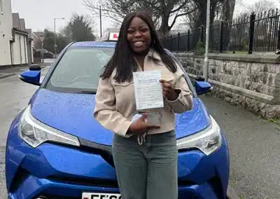 Aminat smiling proudly in front of Fiona's blue automatic driving school car, holding up her practical driving test pass certificate