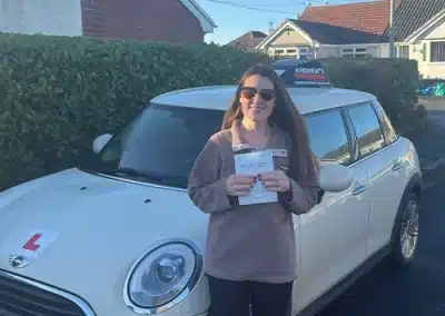 A woman wearing sunglasses and a brown fleece stands in front of a white Mini Cooper learner car, smiling and holding her driving test pass certificate in a residential area of Rhyl.