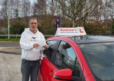 A man named Kevin wearing a white Mustang hoodie, holding his driving test pass certificate next to Claire's red automatic driving school car with an Andrew's Driving roof sign in Colwyn Bay.