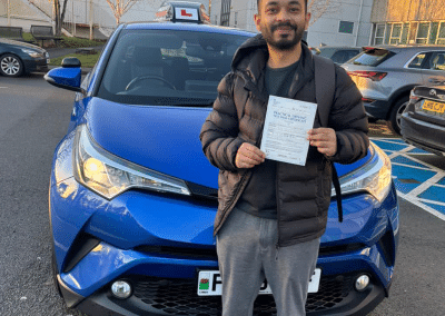 Shah stands next to a blue Toyota C-HR automatic learner car in a parking lot in Rhyl, smiling and holding his driving test pass certificate after a successful lesson with Fiona.