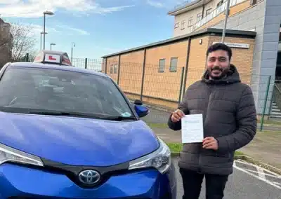 A man in a dark puffer jacket stands next to a blue Toyota C-HR with a learner 'L' sign on top, holding his driving test pass certificate in a parking lot in Rhyl.