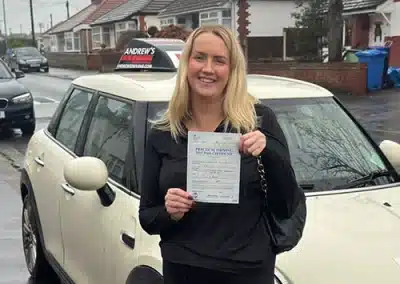 Tasha Burt smiling proudly in front of a white Mini Cooper driving school car, holding her practical driving test pass certificate after passing her automatic driving test with instructor Jimmy Garland