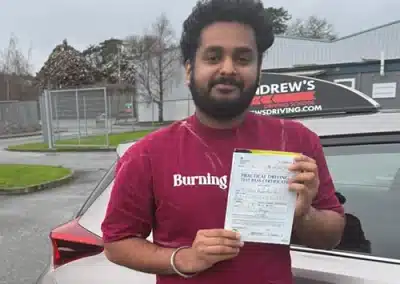 Vishal from Colwyn Bay smiling while holding his practical driving test pass certificate next to an Andrew's Driving School car after lessons with instructor Ursula.