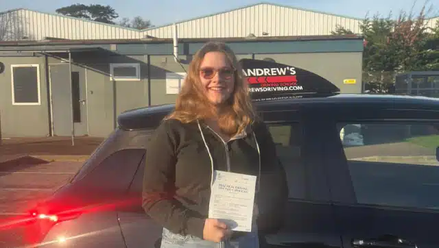 Amelia smiling and holding her driving test pass certificate next to a black car at Bangor Driving Test Centre.