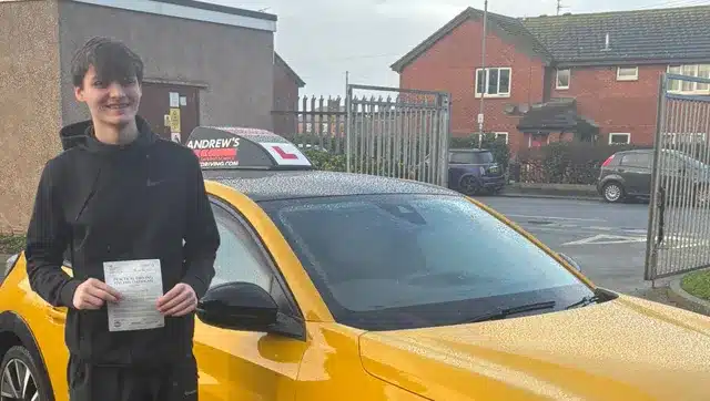 Lloyd holding his driving test pass certificate at Rhyl driving test centre car park beside an Andrew’s learner car.