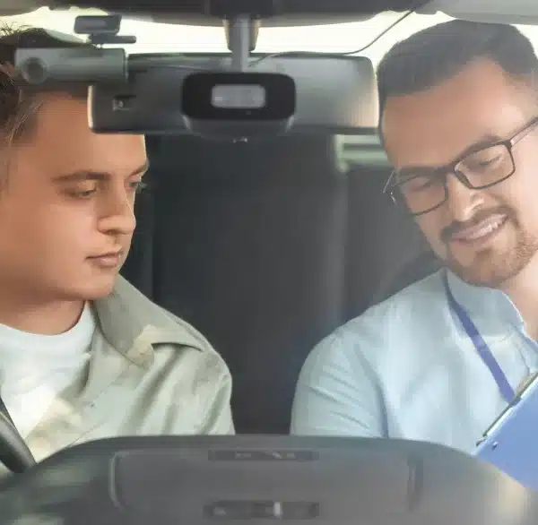 Driving instructor in the front passenger seat assisting a learner driver while reviewing details on a clipboard inside the car.