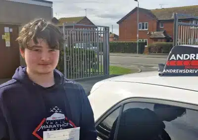 Iori holding a practical driving test pass certificate beside a learner car at Rhyl Driving Test Centre after passing with instructor Paul Sanders.