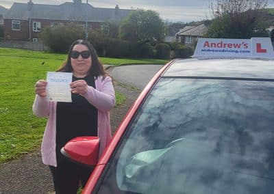 Naz stands on a residential street holding her driving test pass certificate, smiling beside a red learner car with Andrew’s Driving School signage after passing her automatic test in Bangor.