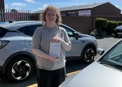 Briana holding her driving test pass certificate at Rhyl Driving Test Centre next to a learner car
