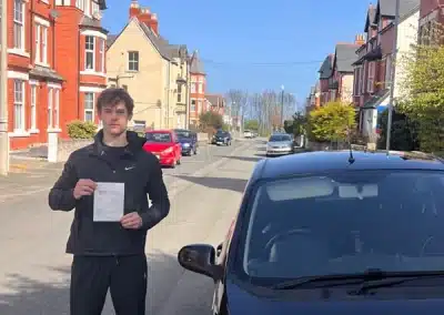 Fred holding his practical driving test pass certificate beside a learner car in Colwyn Bay after passing his test in Rhyl.
