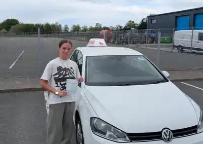 Maisy holding her driving test pass certificate at Bangor Driving Test Centre beside a white learner car