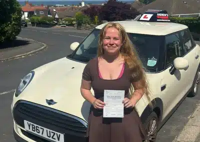 Aimee holding her driving test pass certificate beside an Andrew’s Driving School Mini after passing her automatic driving test in Rhyl.