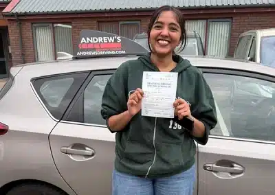 Aleena smiling and holding her driving test pass certificate beside a learner car after passing her driving test in Rhyl with instructor Ursula.