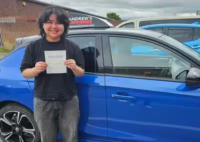 Ashley smiling and proudly holding up a practical driving test pass certificate next to a bright blue Andrew's Driving school car.