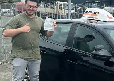 Ashkana holding a driving test pass certificate and giving a thumbs up beside a car after passing first time in Bangor with Geraint.