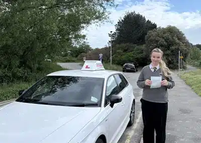 Isabelle standing next to a white Andrew's Driving school car on a sunny day, proudly holding up her practical driving test pass certificate after a first-time pass.