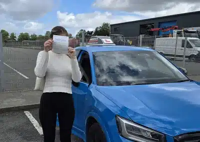 Leah from Colwyn Bay holding her driving test pass certificate beside Ryan's blue car at Bangor Driving Test Centre.