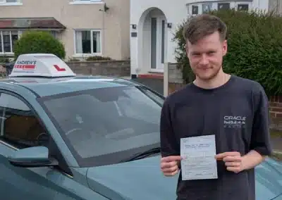 Liam smiling and holding up his driving test pass certificate next to a teal Andrew's Driving school car in a residential area.