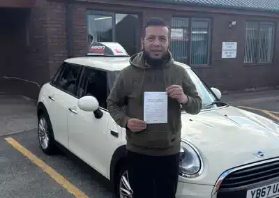 Man standing outside Rhyl Driving Test Centre holding a driving test pass certificate in front of a white Mini with L plates