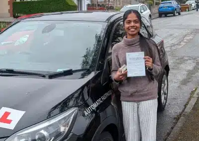 Mihiri stands beside a black automatic learner car in Old Colwyn, holding her driving test pass certificate after passing in Rhyl.