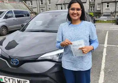 Rejvin standing beside a grey automatic car in London, holding her driving test pass certificate after learning in Rhyl with Fiona.