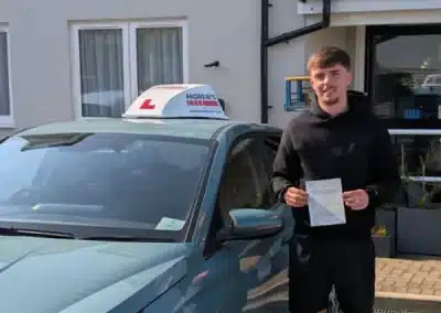 Sam standing beside a learner car outside a modern house in Llandudno Junction, holding his driving pass certificate after passing first time with instructor Jack Nettleton.
