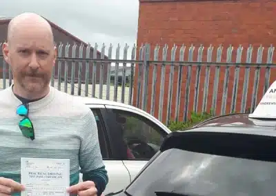 Simon standing in front of a driving school car, proudly holding up his practical driving test pass certificate.