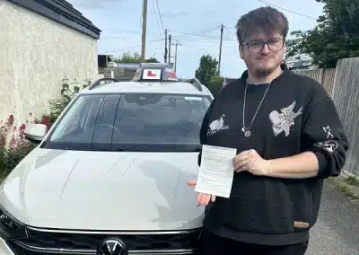 Toby standing beside a white driving school car in Rhyl, holding his driving test pass certificate.