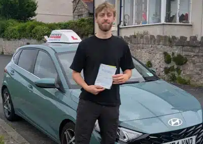Young man holding a driving test pass certificate while standing in front of a car with an L plate on a residential street.