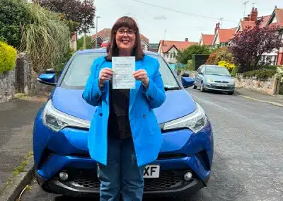 Lindsay proudly holds her driving test pass certificate in front of Fiona’s blue learner car after passing at Rhyl Driving Test Centre.