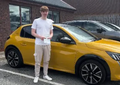 Rylie holding his driving test pass certificate beside a yellow car outside Rhyl Driving Test Centre after passing his driving test.