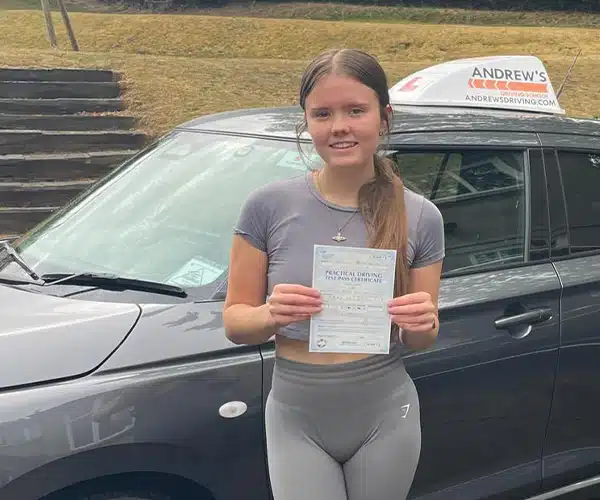 Evelyn Kirkham holding her practical driving test pass certificate beside Andrew’s Driving School car after passing her driving test in Abergele with instructor Andy Kirkham.