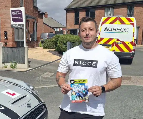 Sam Bailey holding his pass certificate standing in Barnsley holding up his new automatic driving test pass certificate next to his instructor Janets electric Mini