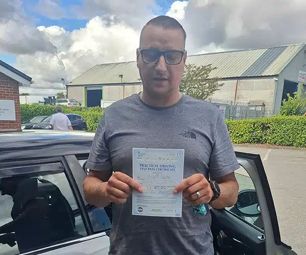 Anthony holding his driving test pass certificate beside Janet’s electric MINI at Barnsley Driving Test Centre.