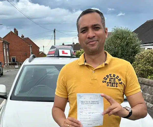 Dr Patel holding his pass certificate standing in front of Paul Singletons car after passing his driving test in Rhyl with Andrew's Driving School.
