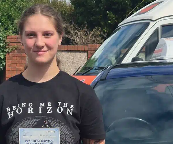 Leah Templeman holding her driving test pass certificate in Wrexham Driving Test Centre beside Craig’s blue driving school car after passing her driving test..