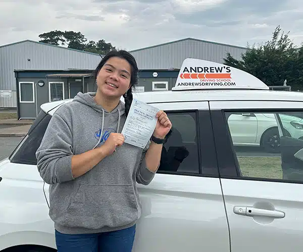 Proy holding her driving test pass certificate, standing by her instructor Robin's white Suzuki Swift in Bangor Driving Test Centre