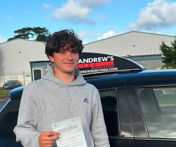 Dwylan outside Bangor Driving Test Centre holding his new driving test pass certificate in front of Llinos's black Suzuki Swift