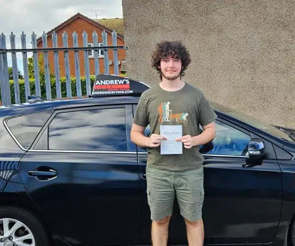 Alfie Baguley holding his pass certificate beside an Andrew’s Driving School car after passing at Rhyl Test Centre