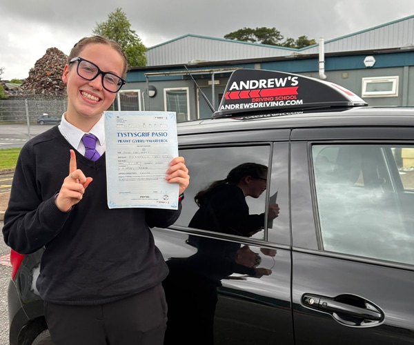 Elliw celebrating after passing her driving test at Bangor Driving Test Centre, proudly holding her Welsh driving test pass certificate beside her Andrew’s Driving School car.