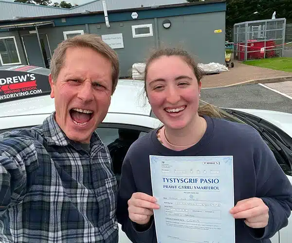 Manon celebrating a first-time driving test pass at Bangor Driving Test Centre with instructor John, proudly holding her Welsh pass certificate beside the Andrew’s Driving School car.