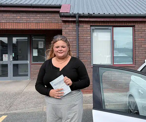 Molly outside Rhyl Driving Test Centre front door holding her test pass cerificate standing next to Marcus's white Audi A3