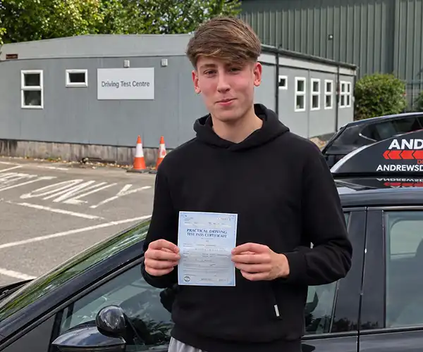 Alfie proudly holding his practical driving test pass certificate beside an Andrew's Driving School learner car, with a driving test centre in the background.