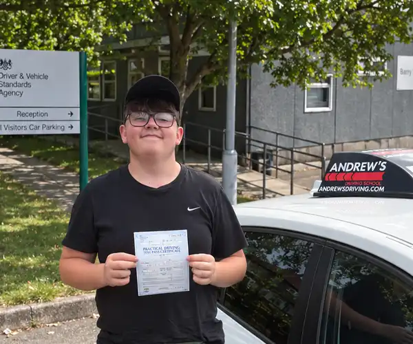 Harry holding his practical driving test pass certificate beside a black Andrew's Driving School tuition car, photographed with a driving test centre building in the background.
