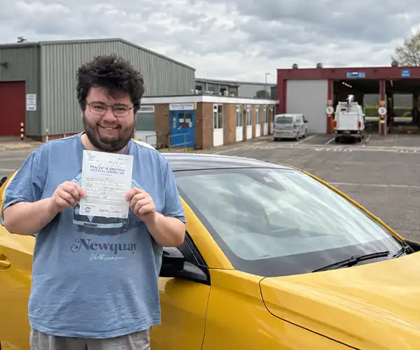 Zoltan smiling and holding his practical driving test pass certificate beside a yellow Peugeot driving tuition car, with a driving test centre in the background.